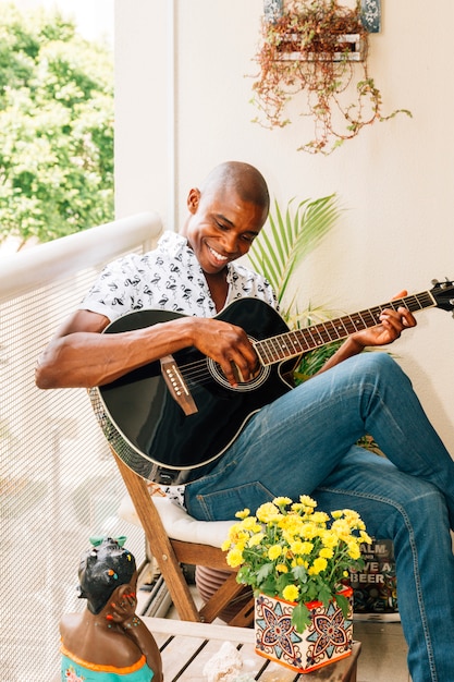 Lächelndes porträt eines afrikanischen jungen mannes, der auf dem stuhl spielt die gitarre im balkon sitzt Kostenlose Fotos