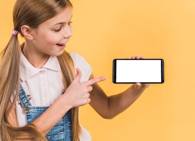 Lächelndes porträt eines mädchens mit dem langen blonden haar zeigend auf den handy, der weißen leeren bildschirm zeigt Kostenlose Fotos