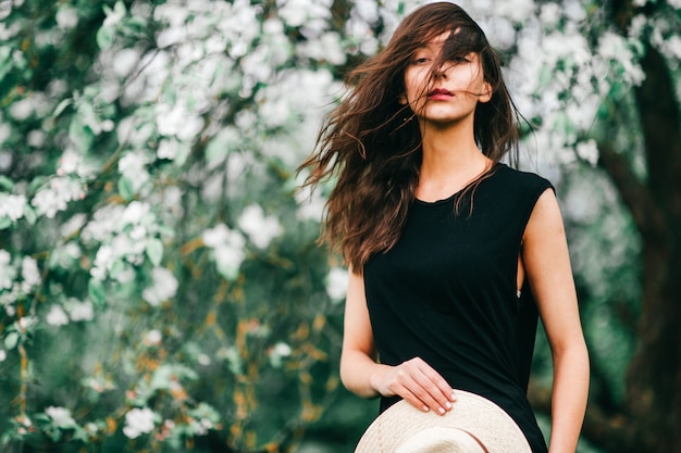 Lebensstilporträt des brunettemädchens im schwarzen kleid mit strohhut in ihren händen über dem blühenden apfelbaum auf hintergrund. Premium Fotos
