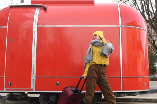 Mädchen läuft mit einem roten koffer. großer retro-oldtimer. altes auto. reisen im winter. mädchen in einem gelben hellen hut und in einer gestrickten strickjacke. reise concept.copy raum Premium Fotos