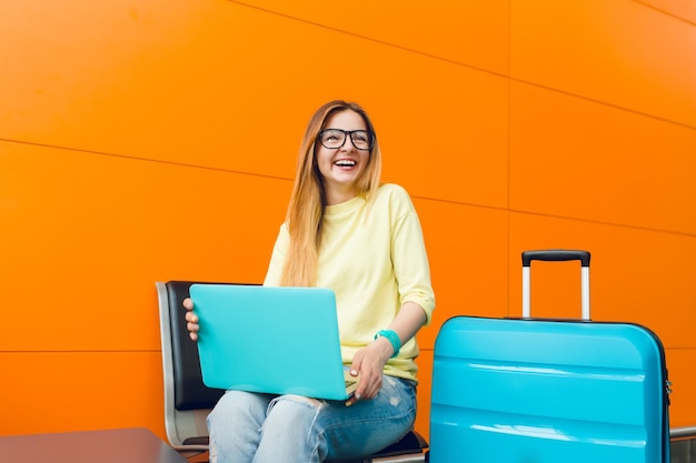 Kostenlose Foto Madchen Mit Langen Haaren Im Gelben Pullover Sitzt Auf Orange Hintergrund Sie Hat Einen Blauen Koffer Und Einen Laptop Sie Lachelt Glucklich