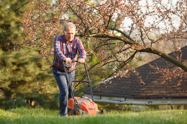 Mann mit elektrischem rasenmäher, rasenmähen. gärtner, der einen garten trimmt. sonniger tag, vorort, dorf. erwachsener mann, der garten beschneidet und landschaftlich gestaltet, gras, rasen, wege trimmend. harte arbeit an der natur. Premium Fotos