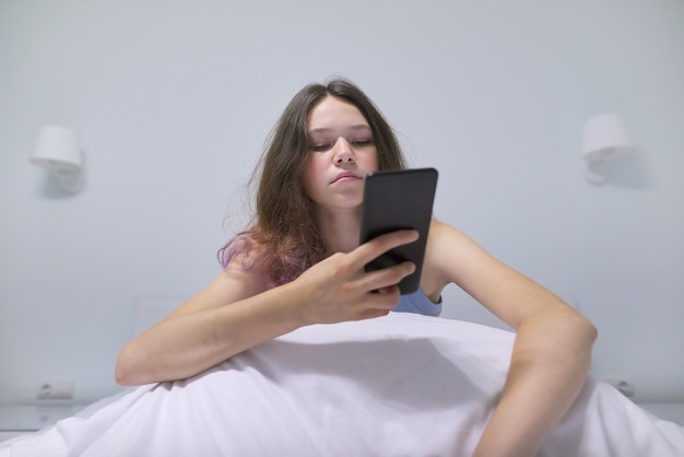 Müder teenager, der zu hause im bett sitzt und handy