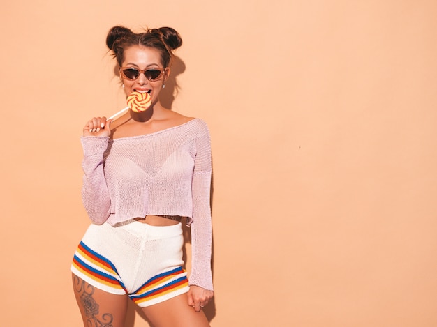 Nahaufnahmeporträt der jungen schönen sexy lächelnden frau mit ghulfrisur. modisches mädchen in der zufälligen sommerkleidung in der sonnenbrille heißes modell lokalisiert auf beige essen, süßigkeitslutscher beißend Kostenlose Fotos