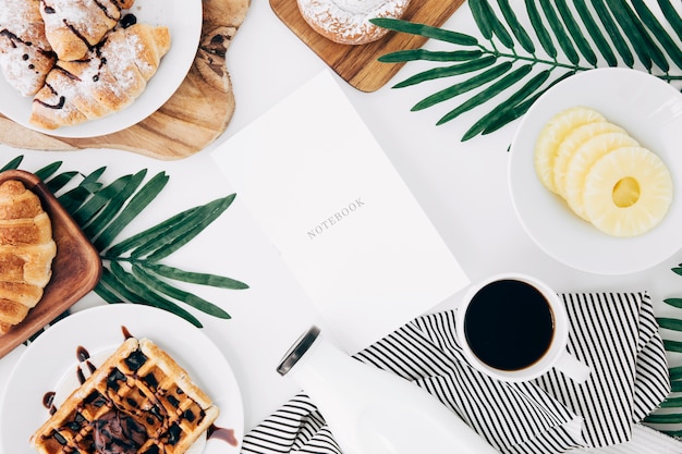 Notizbuch mit croissant umgeben; waffeln; ananasscheiben; flasche und kaffeetasse auf weißem hintergrund Kostenlose Fotos