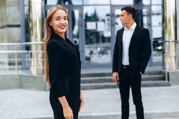 Porträt der glücklichen frau in einer klage, die auf hintergrund des bürogebäudes aufwirft. mann auf hintergrund - bild Premium Fotos