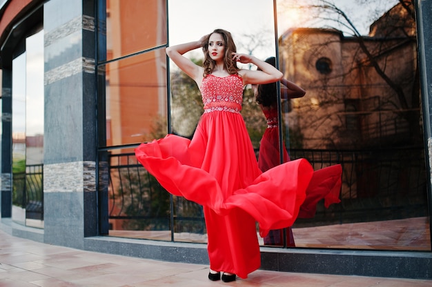 Porträt des modernen mädchens am roten abendkleid warf hintergrundspiegelfenster des modernen gebäudes auf. kleid in die luft blasen Premium Fotos