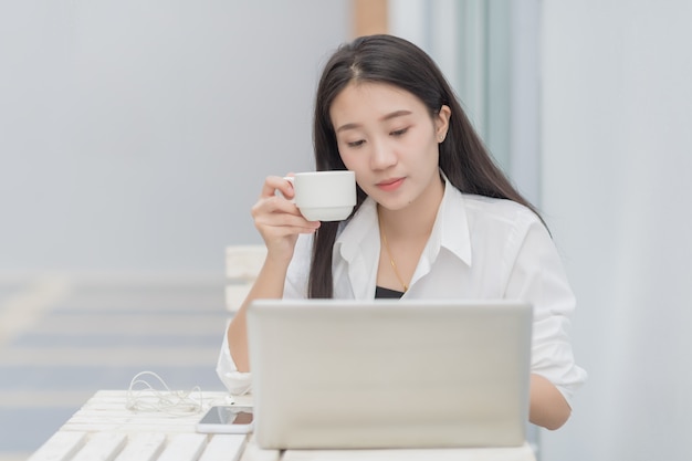 Porträt des netten asiatischen weiblichen modells benutzt laptop-computer für on-line-kommunikation; glücklicher geschäftsfrau-getränkkaffee, der am weißen schreibtisch sitzt. Premium Fotos