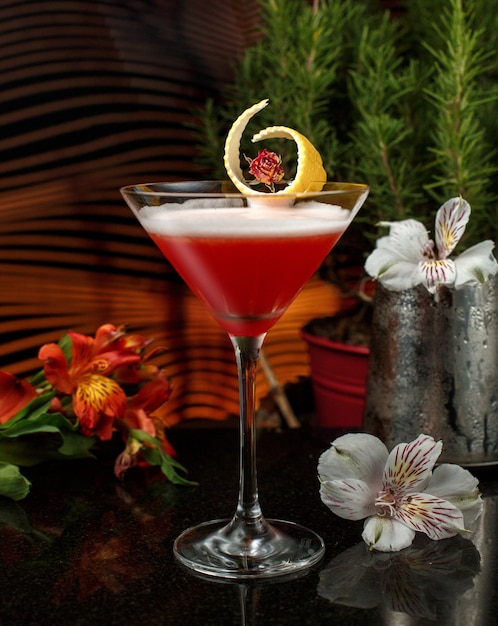 Rotes getränk in martini-glas mit zitronenschale in einer schwach beleuchteten bar mit blumen garnieren Kostenlose Fotos