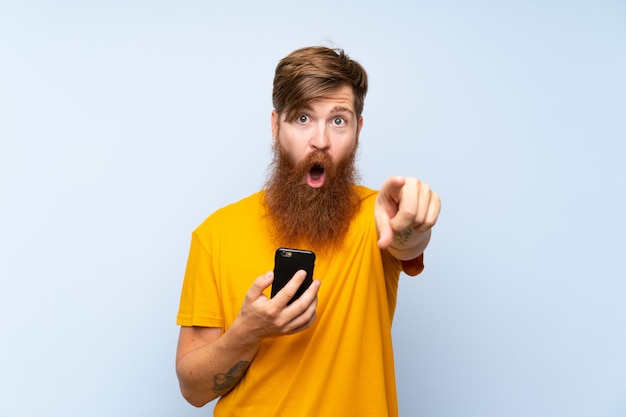 Rothaarigemann mit langem bart mit einem mobile über der blauen wand überrascht und front zeigend Premium Fotos