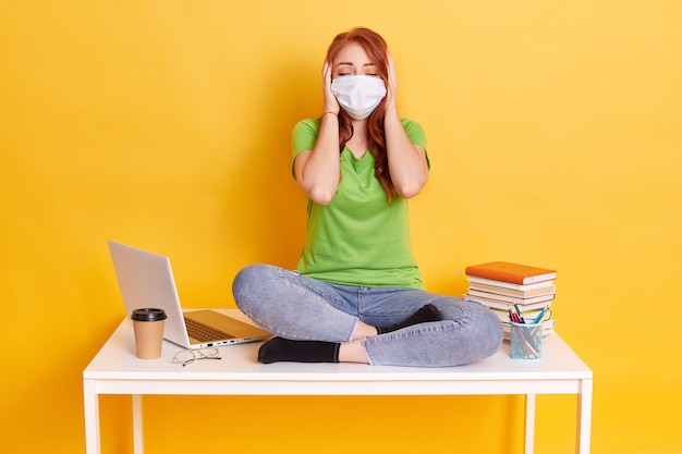 Kostenlos Foto Rothaariges Madchen In Medizinischer Maske Sitzt Mit Gekreuzten Beinen Auf Weissem Tisch Bedeckt Ihre Ohren Mit Handflachen Und Halt Die Augen Geschlossen Tragt Jeans Und T Shirt Umgeben Von Laptop Kaffee