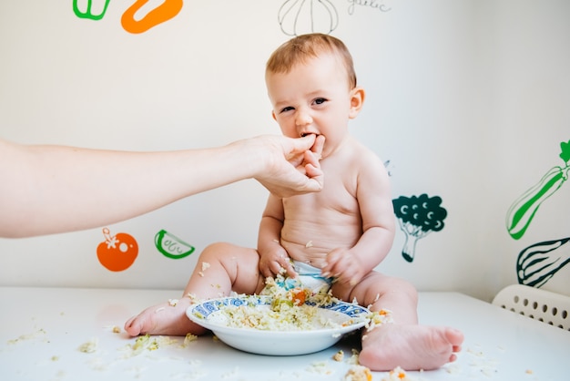 Schmutziges und lächelndes baby auf einer weißen tabelle, die durch die hand seiner mutter eingezogen wird, beim lachen beim versuchen der blw methode. Premium Fotos