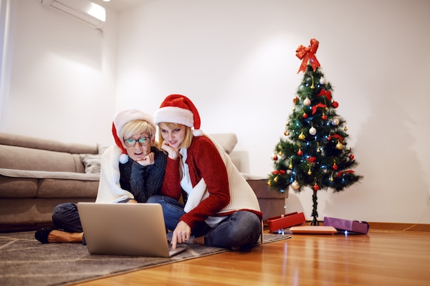 Schone Kaukasische Blonde Frau Die Mit Ihrer Mutter Auf Boden Im Wohnzimmer Sitzt Und Laptop Verwendet Beide Haben Weihnachtsmutzen Auf Den Kopfen Im Hintergrund Ist Weihnachtsbaum Geschmuckt Premium Foto