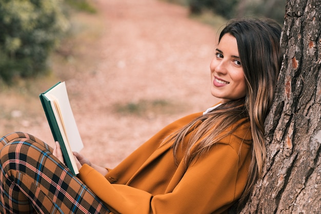 Seitenansicht einer lächelnden jungen frau, die in der hand unter dem baum hält das buch betrachtet kamera sitzt Kostenlose Fotos