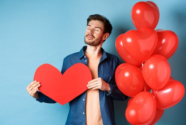 Valentinstag Und Liebeskonzept Vertraumter Mann Mit Geschlossenen Augen Der Romantischen Roten Herzausschnitt Halt Und Nahe Herzballons Blauer Hintergrund Steht Premium Foto