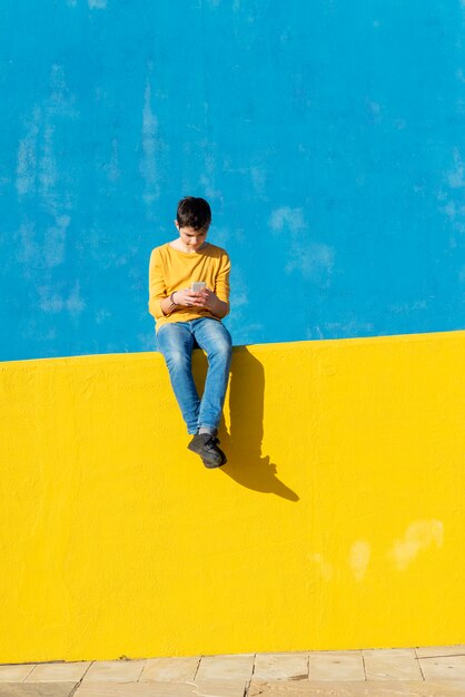 Vorderansicht eines jungen, der die zufällige kleidung sitzt auf einem gelben zaun gegen eine blaue wand bei der anwendung eines smartphone trägt Premium Fotos