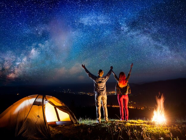Wanderer hoben ihre hände unter den sternen in der nähe von lagerfeuer und zelt auf und blickten nachts auf den sternenhimmel Premium Fotos