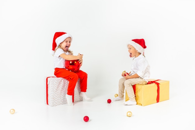 Zwei lustige kleinkinder in sankt-hut, der auf geschenkboxen sitzt. isoliert auf weißem hintergrund weihnachten und neujahr konzept. Premium Fotos