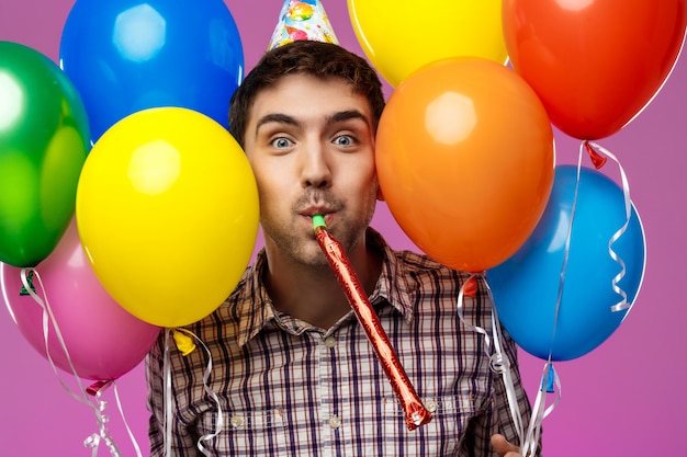 Молодой человек празднует день рождения, держа красочные шарики над фиолетовой стеной. | Бесплатно Фото