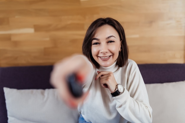 ブルネットの髪を持つ笑顔の女性は彼女のモダンな明るいフラットでテレビをオンにします 無料の写真