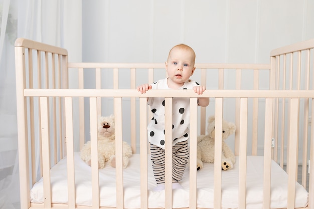 8ヶ月の赤ちゃんが明るい子供部屋のパジャマを着たおもちゃのベビーベッドに立ってカメラを見る プレミアム写真