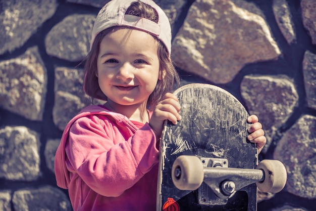 おしゃれな女の子がスケートボードを持って外で遊んでいる 子供の美しい感情 無料の写真