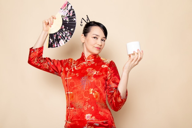 正面の美しい日本の芸者は伝統的な赤い和服でポーズをとって髪棒ポーズ持株扇子とクリーム色の背景にエレガントなクリーム 無料の写真