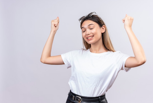 白い壁に目を閉じて握りこぶしの腕を上げる彼女の頭にサングラスをかけている白いtシャツの幸せなかなり若い女性 無料の写真