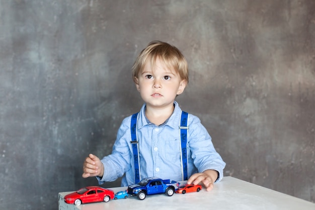 サスペンダー付きのシャツを着た小さな男の子が おもちゃのマルチカラーのおもちゃの車で遊ぶ 自宅のテーブルや保育園でおもちゃの車で遊ぶ幼児男の子 就学前および幼稚園児向けの教育玩具 プレミアム写真