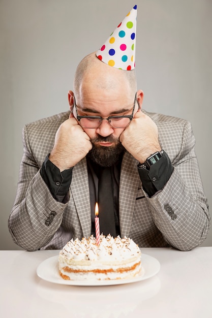 帽子をかぶった一人の男がキャンドルの前でケーキの前に座っています 悲しい誕生日 灰色の壁 垂直 プレミアム写真