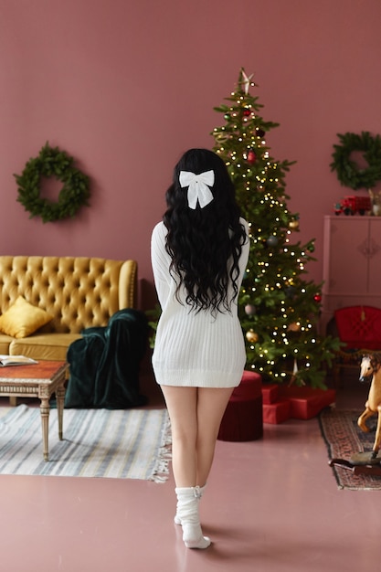 リビングルームのインテリアでクリスマスツリーの近くに彼女の背中とポーズをとって居心地の良いセーターのモデルの女の子 プレミアム写真