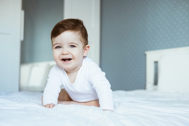 ベッドに座って笑っている白いbodyuitのかわいい幼児の男の子の肖像画 幸せな赤ちゃんの顔は 自宅の寝室の側面図で カメラを見てください プレミアム写真