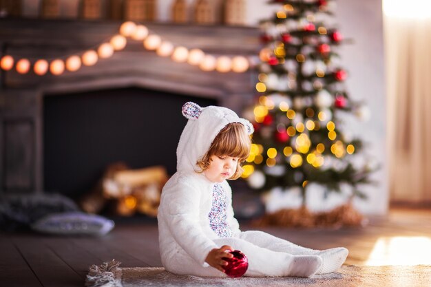 白いウサギの衣装を着た小さな子供がクリスマスツリーの近くに座って 赤いクリスマスボールを持っています 高品質の写真 プレミアム写真
