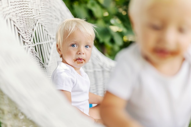 青い目と汚れたチョコレートの顔を持つブロンドの髪の幼児は 庭の籐の椅子に座ってカメラを見ています かわいい赤ちゃんはお菓子を楽しんでいます 赤ちゃんの 面白い写真 プレミアム写真