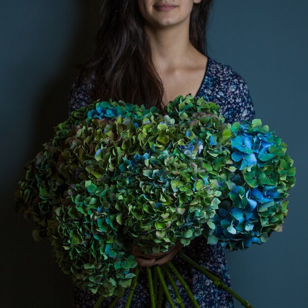 部屋の壁に手で装飾的な緑と青の葉の花の花束を保持している女性 無料の写真