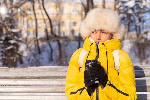 晴れた日に雪に覆われた公園で暖かい服を着た冬の女性はベンチに座って寒さから凍っていて 冬は不幸で 一人でコーヒーを持っています 無料の写真