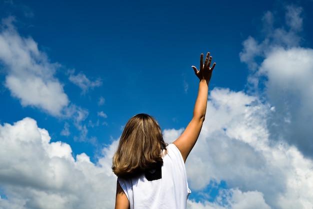 白いtシャツを着て 雲のある青い空を背景に 写真家に背を向けて立っている女性 空に手を伸ばす プレミアム写真