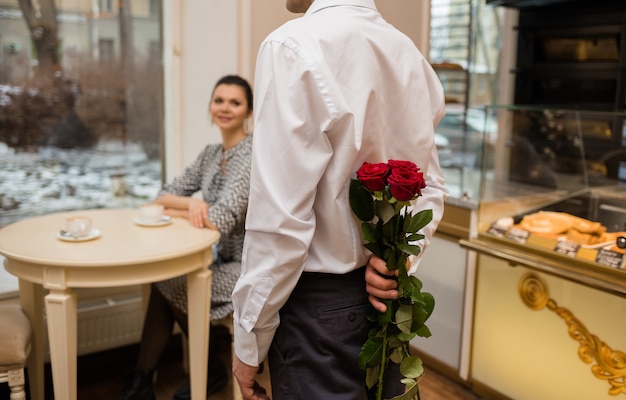 シャツを着た若い男が背中の後ろにバラの花束を持っています カフェでデート プレミアム写真