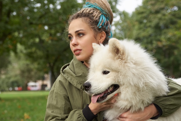 緑のコートを着て 頭にドレッドヘアを付け 公園でサモエド犬と一緒に休む若い女性 プレミアム写真