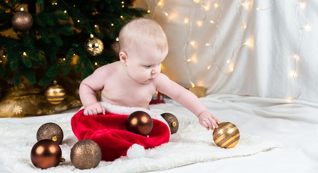 Premium Photo | Adorable baby withoun clothing on santa claus hat on a  background of christmas balls