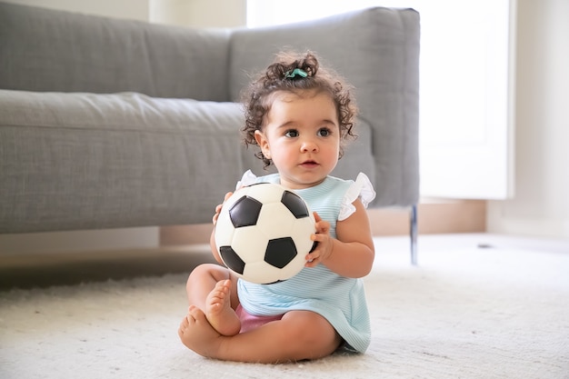 家の床に座って 目をそらし サッカーボールをしている淡いブルーの服を着た愛らしい黒髪の女の赤ちゃん 家庭での子供と子供の頃のコンセプト 無料の写真