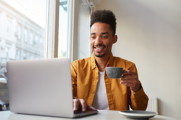 カフェのテーブルに座ってノートパソコンで作業しているアフリカ系アメリカ人の男は 黄色いシャツを着て 香りのよいコーヒーを飲み ガールフレンドとおしゃべりをして 一日を楽しんでいます 無料の写真