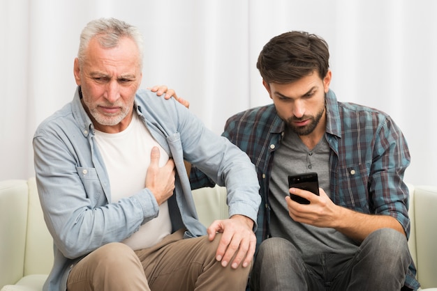 Aged man having heart attack near young guy using smartphone on sofa Free Photo