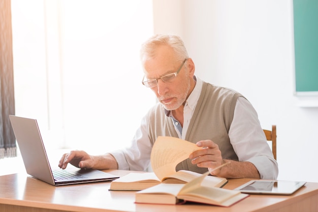 教室で本を読みながらノートパソコンを扱う高齢者教授男性 無料の写真