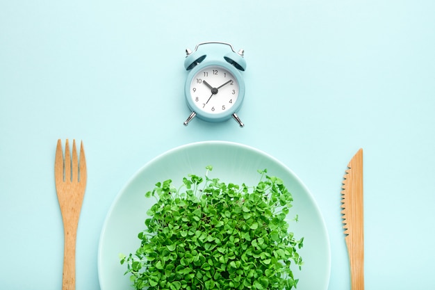 Alarm clock, cutlery and plate with greenery on blue. Premium Photo