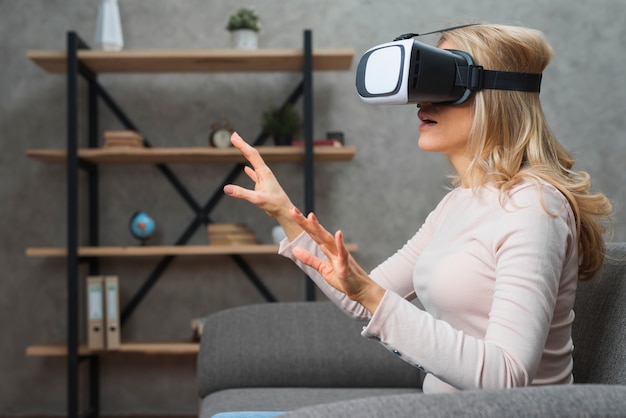 Amazed young woman sitting on sofa in 3d goggles trying to touch something invisible in the air