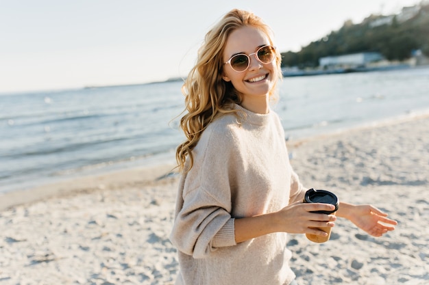 ビーチで一杯のコーヒーを保持している素晴らしい盲目の女性 寒い日に湖の近くでポーズをとるサングラスの熱狂的な女性モデル 無料の写真