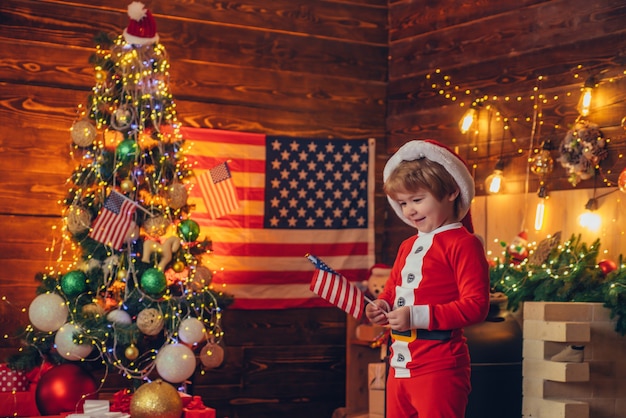 アメリカの伝統の概念 かわいい赤ちゃんの幼児はクリスマスを祝います 旗を振っているアメリカの子供陽気な気分 子供はアメリカの国旗のクリスマスツリーを再生します 楽しんでいる小さな男の子のサンタの帽子と衣装 プレミアム写真