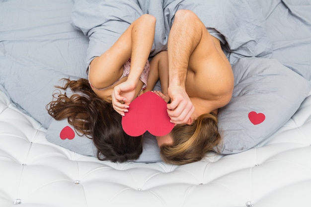彼らの顔の上にハート形の紙を保持しているベッドで寝ている若いカップルの俯瞰 無料の写真