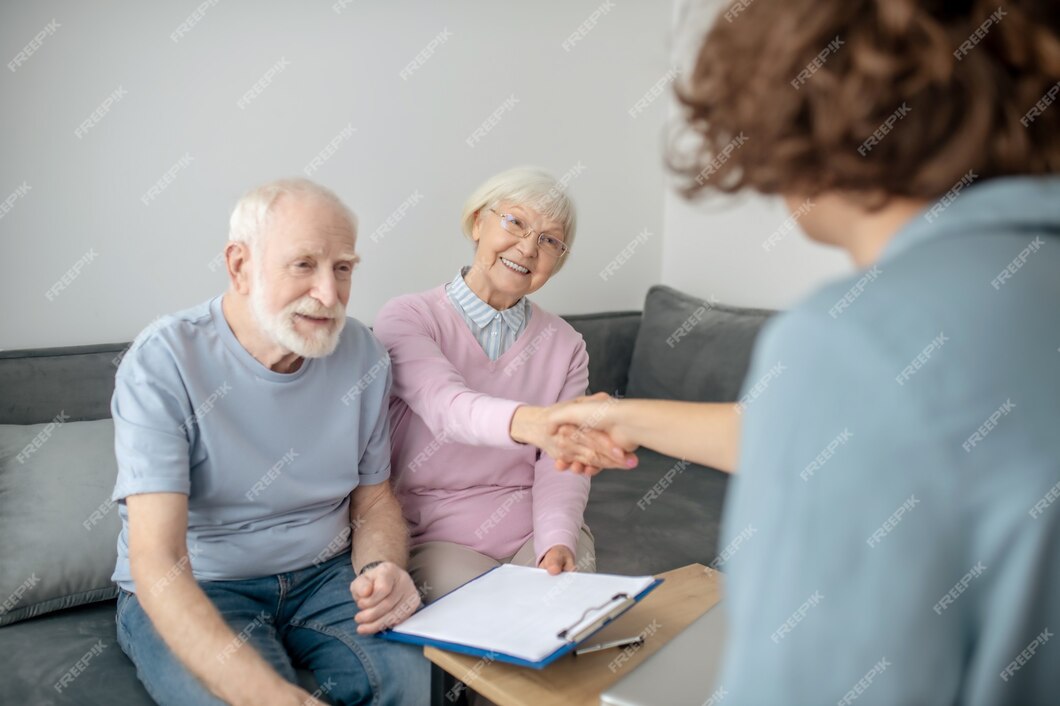  A senior couple is sitting on a couch in a living room, talking to a life insurance agent about whole life insurance.